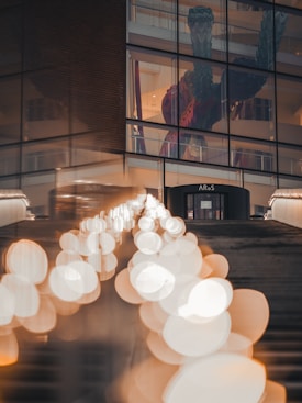 A modern art museum building with large glass windows, through which a giant sculpture of a humanoid figure with a colorful outfit is visible. The foreground features a staircase with bright circular bokeh lights creating a dreamy, out-of-focus effect.