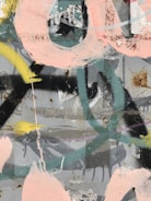 A textured surface featuring overlapping layers of spray paint and brush strokes in an abstract design. The image includes a mix of pastel pinks, yellows, and greens against a backdrop of gray and black. There are signs of weathering and rust on the surface, adding character to the artwork.