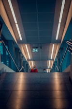 An indoor staircase view with two people visible at the top, both wearing face masks. The ceiling is illuminated with modern LED lights, and there's a green emergency exit sign. The stairs are bordered by sleek metal railings, creating a modern architectural feel.
