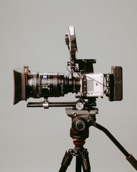 A professional video camera mounted on a tripod. The camera has various attachments, including a large lens, a viewfinder, and other equipment, suggesting it is set up for high-quality filming. The background is a neutral gray color, highlighting the details of the camera.