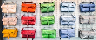 A display of colorful handbags in three rows on a perforated white wall, each row showcasing bags in different shades such as orange, green, blue, red, yellow, and beige.