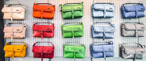 A display of colorful handbags in three rows on a perforated white wall, each row showcasing bags in different shades such as orange, green, blue, red, yellow, and beige.