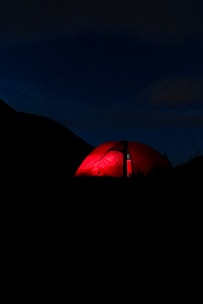 A brightly lit red tent is illuminated against a dark, night backdrop. The surrounding area is mostly shrouded in darkness, creating a stark contrast with the vivid tent.