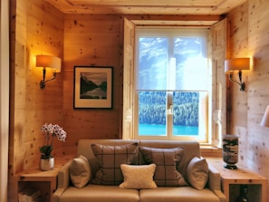 A cozy room featuring wooden walls and warm lighting from two wall-mounted lamps. A comfortable sofa with decorative cushions is centered below a window showcasing a stunning outdoor view of a lake surrounded by trees and mountains. A small wooden side table holds a potted plant with purple flowers on one side and a decorative vase on the other. There is also a framed artwork hanging on the wall.