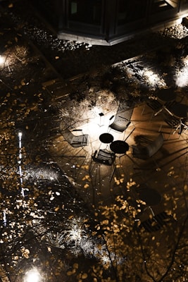 An outdoor seating area viewed from above at night, with multiple metal chairs and tables arranged on a wooden deck. Soft lighting illuminates the patio, creating shadows on the ground. Surrounding the area are decorative grasses and small bushes, with some stone or gravel landscaping.