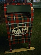 A decorative mailbox is adorned with a red and green plaid pattern and is labeled 'North Pole Airmail Express Service'. It is situated outdoors on a grass surface.