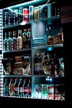 A vending machine filled with a variety of drinks and snacks. The upper shelves contain cans of soft drinks and energy drinks, while the middle section displays bottles of beverages and packaged snacks like chips and nuts. The lower shelves hold bottled sodas and more snacks. The vending machine is illuminated with bright LED lights, highlighting the colorful packaging.