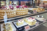 An assortment of cakes and pastries is displayed behind a glass case. At the top, there are tiered cakes adorned with white cream dollops and bright purple flowers. Next to them, lemon meringue pies are arranged neatly. The bottom shelf showcases a variety of cheesecakes and tiramisu, decorated with cream and raspberries. Each item has a label with its name and price.