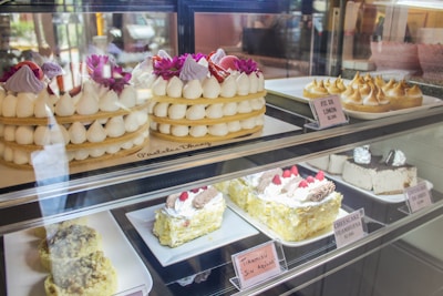 An assortment of cakes and pastries is displayed behind a glass case. At the top, there are tiered cakes adorned with white cream dollops and bright purple flowers. Next to them, lemon meringue pies are arranged neatly. The bottom shelf showcases a variety of cheesecakes and tiramisu, decorated with cream and raspberries. Each item has a label with its name and price.