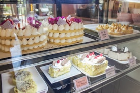 An assortment of cakes and pastries is displayed behind a glass case. At the top, there are tiered cakes adorned with white cream dollops and bright purple flowers. Next to them, lemon meringue pies are arranged neatly. The bottom shelf showcases a variety of cheesecakes and tiramisu, decorated with cream and raspberries. Each item has a label with its name and price.