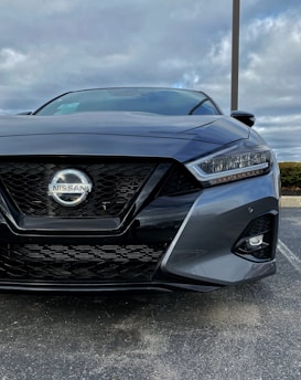A close-up view of the front of a dark-colored Nissan car with modern headlight design and a prominent grille featuring the Nissan logo. The sky is overcast, creating a subdued lighting effect, with a parking lot visible in the background.
