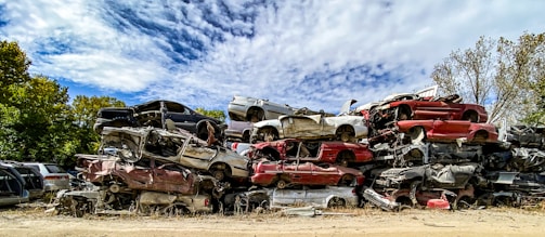 Several stacks of crushed cars in various colors are piled together in an open area, surrounded by trees under a partially cloudy sky.