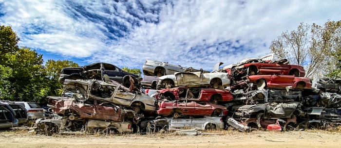 Several stacks of crushed cars in various colors are piled together in an open area, surrounded by trees under a partially cloudy sky.