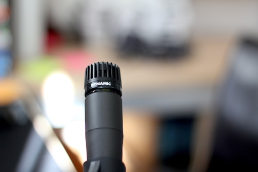 A black microphone is prominently featured in the foreground with the word 'DYNAMIC' printed on its mesh grille. The background is blurred, displaying muted colors and indistinct shapes, suggesting an indoor setting.