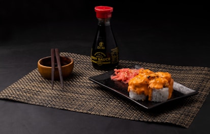 A neatly arranged sushi meal featuring rolls topped with creamy orange sauce on a black square plate. Pickled ginger is placed alongside the sushi. A bottle of soy sauce with a red cap is positioned in the background. Wooden chopsticks rest on a small wooden bowl, all placed on a woven placemat.