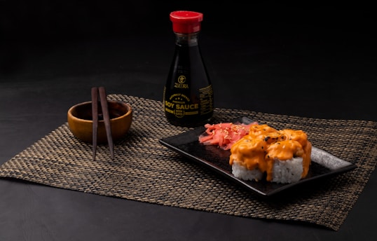 A neatly arranged sushi meal featuring rolls topped with creamy orange sauce on a black square plate. Pickled ginger is placed alongside the sushi. A bottle of soy sauce with a red cap is positioned in the background. Wooden chopsticks rest on a small wooden bowl, all placed on a woven placemat.