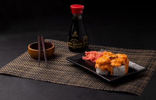 A neatly arranged sushi meal featuring rolls topped with creamy orange sauce on a black square plate. Pickled ginger is placed alongside the sushi. A bottle of soy sauce with a red cap is positioned in the background. Wooden chopsticks rest on a small wooden bowl, all placed on a woven placemat.