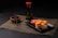 A neatly arranged sushi meal featuring rolls topped with creamy orange sauce on a black square plate. Pickled ginger is placed alongside the sushi. A bottle of soy sauce with a red cap is positioned in the background. Wooden chopsticks rest on a small wooden bowl, all placed on a woven placemat.