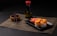 A neatly arranged sushi meal featuring rolls topped with creamy orange sauce on a black square plate. Pickled ginger is placed alongside the sushi. A bottle of soy sauce with a red cap is positioned in the background. Wooden chopsticks rest on a small wooden bowl, all placed on a woven placemat.