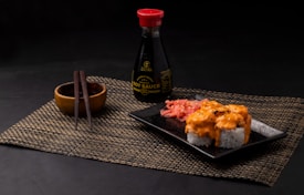 A neatly arranged sushi meal featuring rolls topped with creamy orange sauce on a black square plate. Pickled ginger is placed alongside the sushi. A bottle of soy sauce with a red cap is positioned in the background. Wooden chopsticks rest on a small wooden bowl, all placed on a woven placemat.