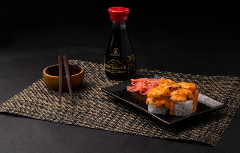 A neatly arranged sushi meal featuring rolls topped with creamy orange sauce on a black square plate. Pickled ginger is placed alongside the sushi. A bottle of soy sauce with a red cap is positioned in the background. Wooden chopsticks rest on a small wooden bowl, all placed on a woven placemat.