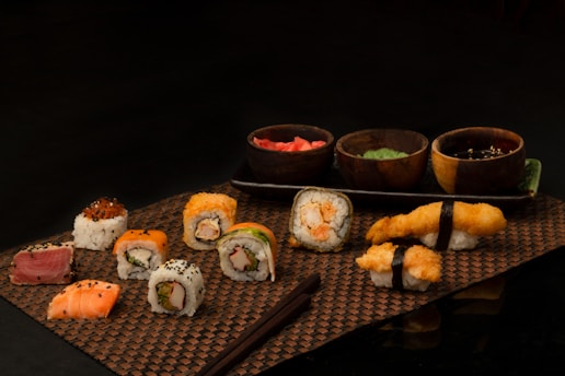 An assortment of sushi rolls and nigiri displayed on a woven mat with wooden bowls filled with different sauces or garnishes. The sushi pieces are diverse, featuring various ingredients like fish, rice, and seaweed. Two pairs of chopsticks rest on the mat beside the sushi.