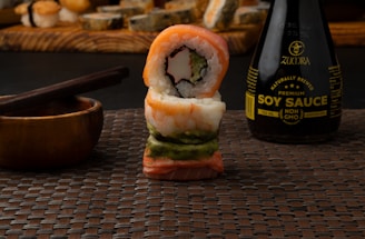 A stack of sushi rolls is placed on a woven mat. The rolls are intricately arranged with various layers, showcasing ingredients like rice, seaweed, fish, and avocado. A wooden bowl with chopsticks rests nearby, and in the background, there is a bottle of soy sauce labeled 'Zumra Premium Soy Sauce.' The background features a blurred assortment of more sushi rolls on a wooden board.