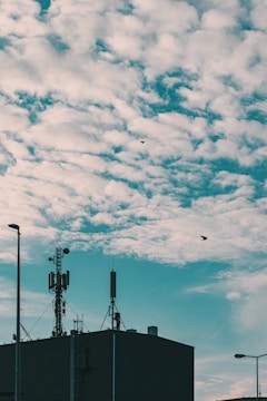 A rooftop with various antennas and communication equipment is silhouetted against a sky filled with textured clouds. Two birds are flying in the sky, adding a sense of movement to the scene. Streetlights are visible on either side of the building.
