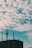 A rooftop with various antennas and communication equipment is silhouetted against a sky filled with textured clouds. Two birds are flying in the sky, adding a sense of movement to the scene. Streetlights are visible on either side of the building.