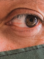 A detailed close-up of a human eye, revealing the intricate patterns and colors within the iris, surrounded by eyelashes. The lower part of the image shows a portion of fabric, possibly a mask, indicating a focus on personal protection or attention to features.