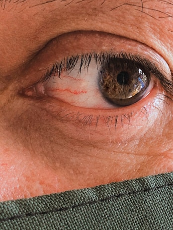 A detailed close-up of a human eye, revealing the intricate patterns and colors within the iris, surrounded by eyelashes. The lower part of the image shows a portion of fabric, possibly a mask, indicating a focus on personal protection or attention to features.