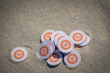 A collection of circular tokens with the Bitcoin logo lies scattered on sand. Each token features an orange circle with a white border and the symbolic Bitcoin 'B' in the center.