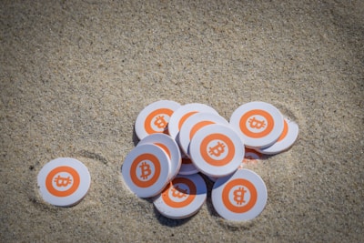A collection of circular tokens with the Bitcoin logo lies scattered on sand. Each token features an orange circle with a white border and the symbolic Bitcoin 'B' in the center.