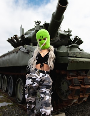 A person with long blonde hair is wearing a bright green ski mask with dollar signs and a black crop top. They are leaning against a large military tank. The tank is green and has visible tracks and a cannon. The person is also wearing camouflage pants and appears to be outdoors under a cloudy sky.