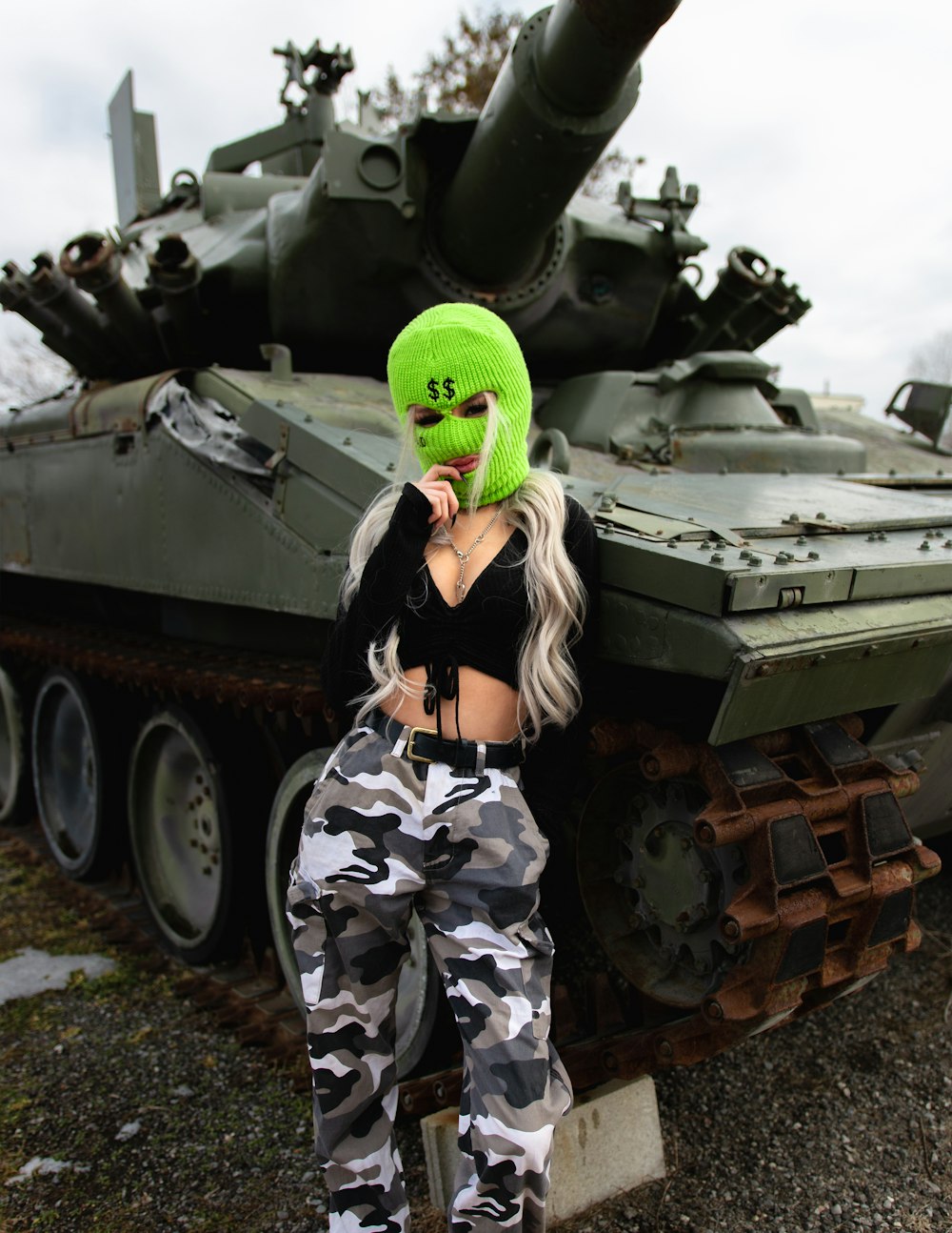 緑と黒の迷彩服を着た女性が緑の戦車のそばに立つ