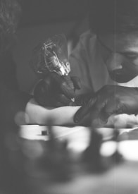 A tattoo artist is intently focused on tattooing a person's arm. The scene is captured in black and white, with soft lighting and a shallow depth of field that blurs the background. The tattoo artist is wearing black gloves and holding a tattoo machine.