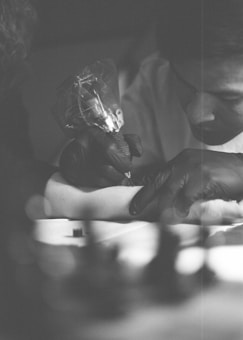 A tattoo artist is intently focused on tattooing a person's arm. The scene is captured in black and white, with soft lighting and a shallow depth of field that blurs the background. The tattoo artist is wearing black gloves and holding a tattoo machine.