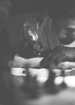 A tattoo artist is intently focused on tattooing a person's arm. The scene is captured in black and white, with soft lighting and a shallow depth of field that blurs the background. The tattoo artist is wearing black gloves and holding a tattoo machine.