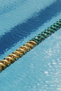 A swimming pool has clear blue water with sunlight reflecting on its surface, creating a shimmering effect. A lane divider floats horizontally across the pool, comprised of alternating yellow and green sections, creating a visually appealing pattern.