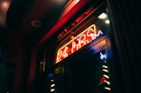 A neon sign in a dark setting with red, blue, and yellow colors displaying '24 HRS'. It casts a warm glow onto a glass surface, creating reflections.