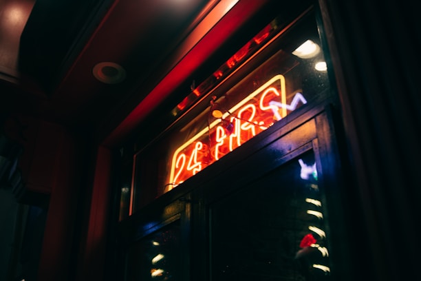 A neon sign in a dark setting with red, blue, and yellow colors displaying '24 HRS'. It casts a warm glow onto a glass surface, creating reflections.