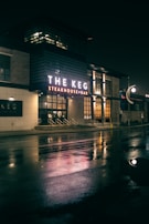 A night scene featuring a steakhouse with illuminated signage that reads 'The Keg Steakhouse + Bar'. The bright lights create reflections on the wet pavement. The building has a modern exterior with brick and glass elements, and there are steps leading to the entrance.