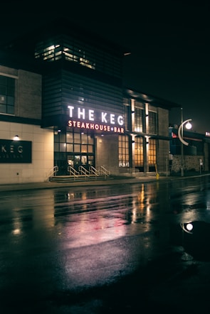 A night scene featuring a steakhouse with illuminated signage that reads 'The Keg Steakhouse + Bar'. The bright lights create reflections on the wet pavement. The building has a modern exterior with brick and glass elements, and there are steps leading to the entrance.