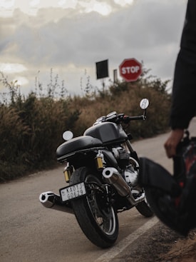 A motorcycle is parked on a rural road, with a visible license plate. A stop sign is positioned on the roadside amidst tall grass and shrubs. The sky is overcast, casting a subdued, moody lighting across the scene. Part of a person's arm and hand holding a helmet can be seen in the foreground.