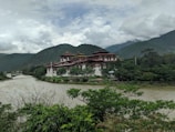 A large, traditional Bhutanese fortress-like building, known as a dzong, is situated on a scenic riverbank. Surrounded by lush greenery, it is set against a backdrop of misty, forested mountains under a partly cloudy sky.