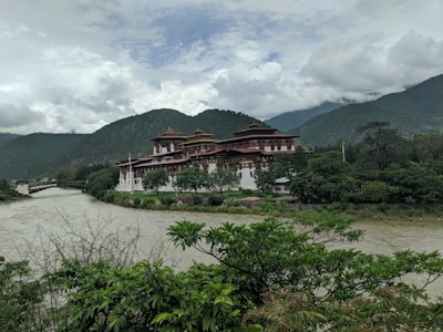 A large, traditional Bhutanese fortress-like building, known as a dzong, is situated on a scenic riverbank. Surrounded by lush greenery, it is set against a backdrop of misty, forested mountains under a partly cloudy sky.