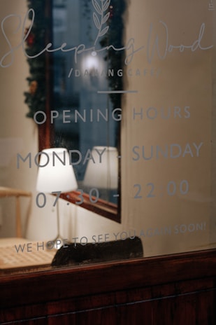 A glass window shows the name of a café 'Sleeping Wood' along with its opening hours, which are Monday to Sunday from 07:30 to 22:00. Inside the café, a wooden table and a lamp are visible, creating a warm and inviting ambiance.