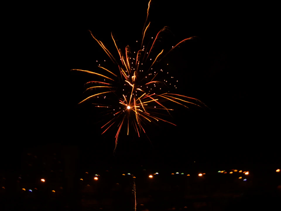 The image depicts a fireworks display against a dark night sky, with orange and yellow sparks illuminating the scene. The fireworks are located centrally in the frame, creating a sense of depth and perspective. In the background, silhouettes of buildings can be seen, suggesting that this is taking place in an urban setting or near a populated area.