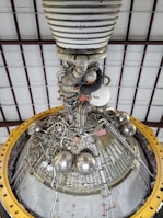 A detailed view of a complex engine or machinery component, featuring numerous metallic pipes, valves, and spherical objects. The structure is surrounded by a large yellow circular frame and is positioned against a ceiling with a grid-like pattern of beams.