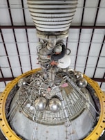 A detailed view of a complex engine or machinery component, featuring numerous metallic pipes, valves, and spherical objects. The structure is surrounded by a large yellow circular frame and is positioned against a ceiling with a grid-like pattern of beams.